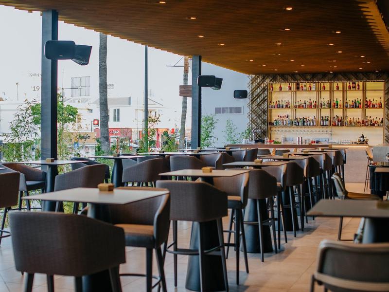 Modernes Café mit großen Fenstern, Holzdecke, schwarzen Stühlen und vielen Tischen.