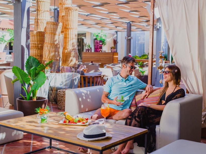 Zwei Personen sitzen entspannt auf grauen Sofas in hellem Café mit Holztisch und Pflanzen.