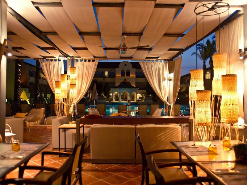 Gemütliche Lounge mit warmem Licht, Holzmöbeln und Blick auf Pool im Abendlicht.