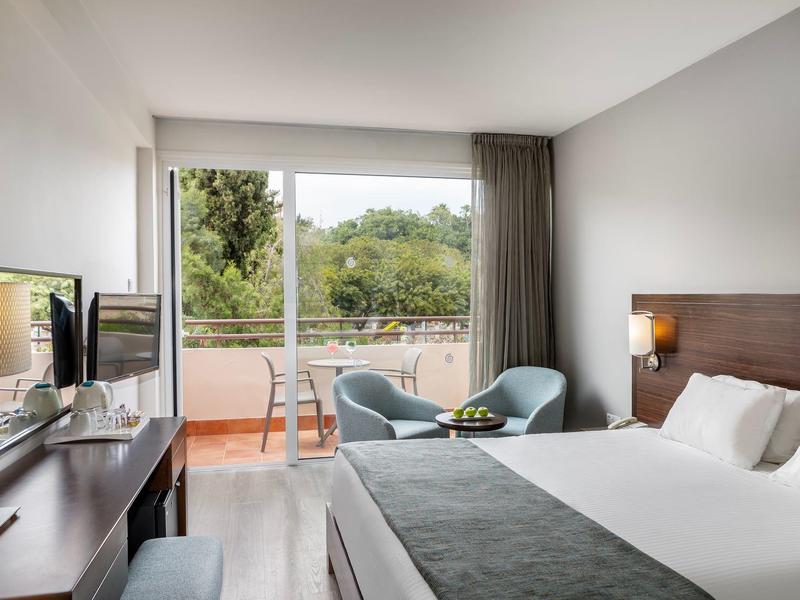 Helles Hotelzimmer mit Doppelbett, Sesseln, Schreibtisch und Balkon mit Blick auf Bäume.