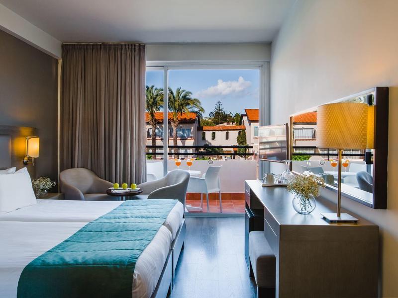 Modernes Hotelzimmer mit zwei Betten, Schreibtisch, Stuhl und Blick auf Pool und Palmen.