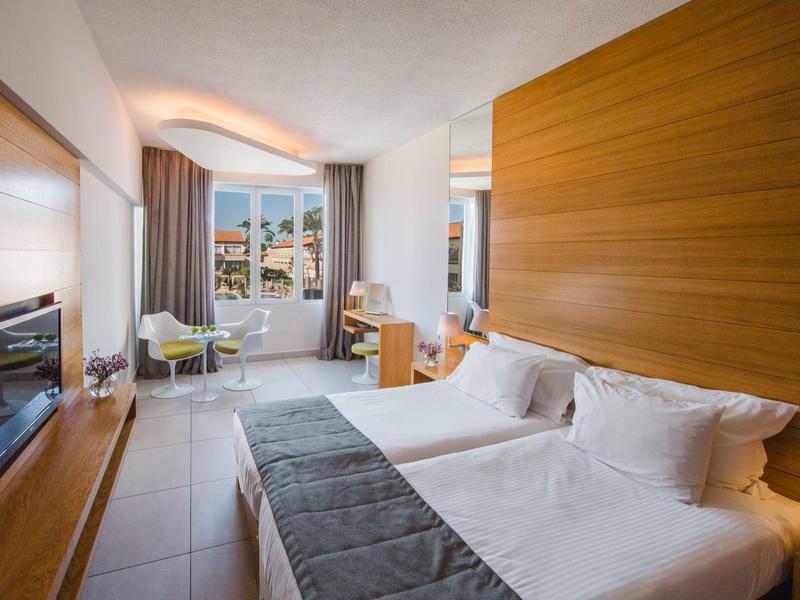 Modernes Hotelzimmer mit zwei Betten, Holzpaneelen, Schreibtisch und Sitzbereich am Fenster.