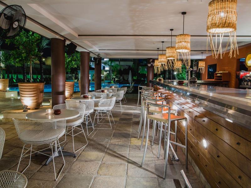 Moderner Außenbereich einer Bar mit weißen Stühlen, Holztresen und warmen Hängeleuchten.