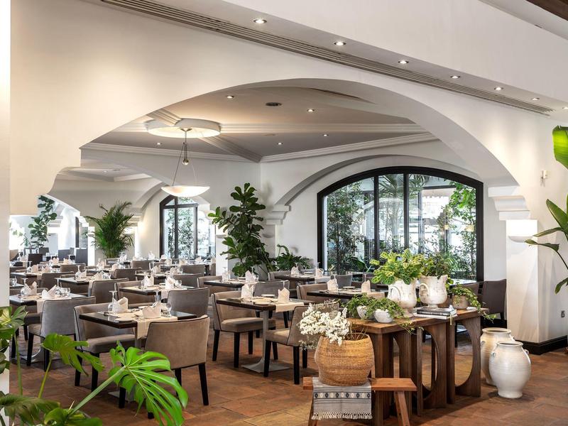 Helles Restaurant mit Holzböden, gebogenen Decken, vielen Pflanzen und gedeckten Tischen.