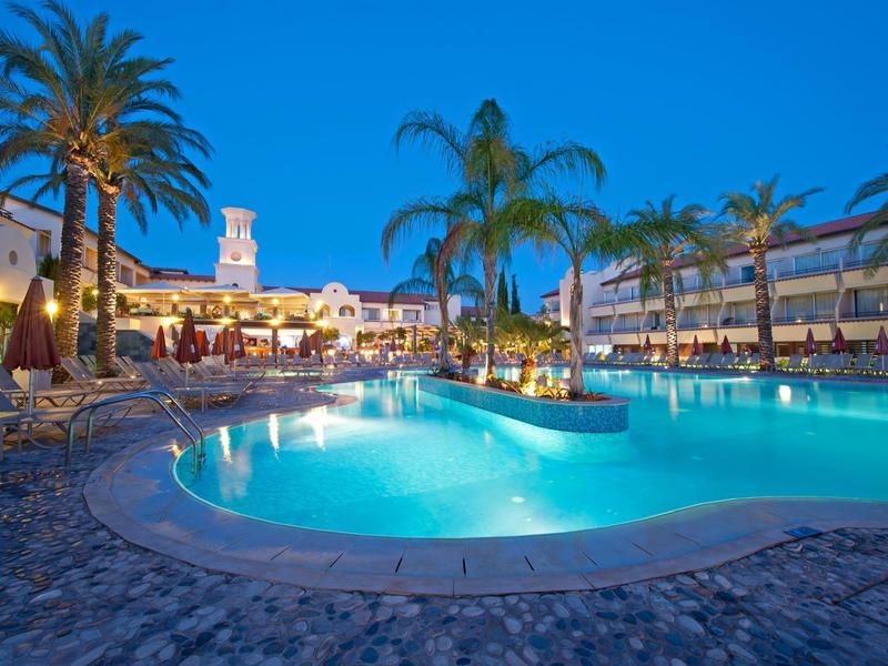 Ein großer beleuchteter Hotelpool mit Palmen und umliegenden Gebäuden bei Dämmerung.