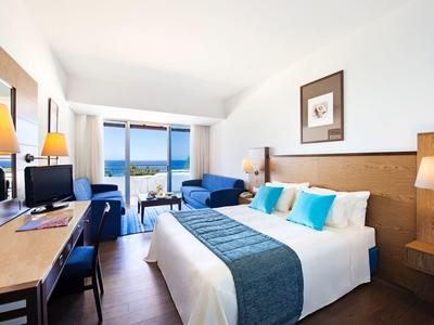 Bright hotel room with large bed, desk, and balcony overlooking the sea.