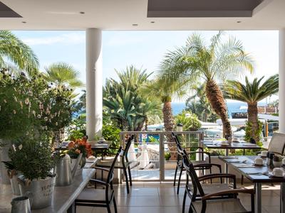 Restaurant terrace with chairs and tables, view of palm trees and sea in the background.