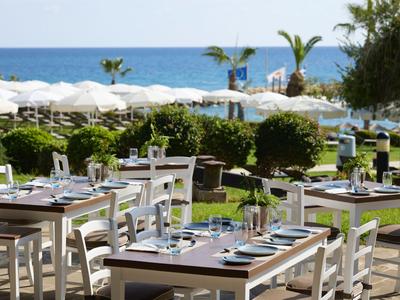 Terrasse eines Restaurants mit weißen Tischen und Stühlen am Meer mit Sonnenschirmen im Hintergrund