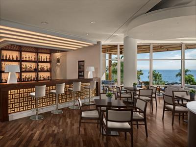 Elegante Hotelbar mit Holzfußboden, Barhockern und großen Fenstern mit Blick auf das Meer.