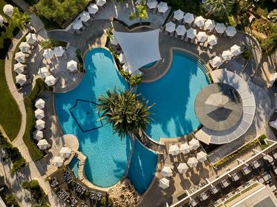 Blick von oben auf einen hotelpool mit geschwungenem design, umgeben von weißen Sonnenschirmen und Liegen.
