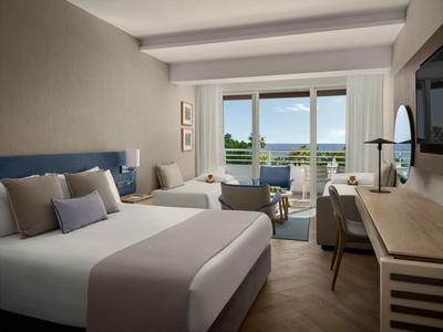 Modernes Hotelzimmer mit zwei Betten, Balkon und Meerblick unter hellem Licht.