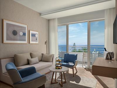 Helles Hotelzimmer mit Sofa, Sesseln, kleinem Tisch und Meerblick durch große Fenster.