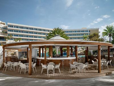 Offener Pavillon mit Sitzgelegenheiten vor einem mehrstöckigen Hotel unter blauem Himmel.