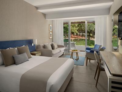 Moderne Hotellounge mit großem Bett, Sofa, Schreibtisch und großen Fenstern mit Blick auf Garten.