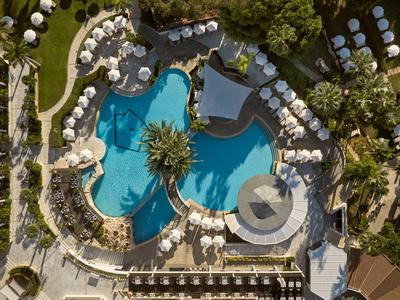 Luftaufnahme eines Hotelpools mit Sonnenschirmen und Liegestühlen in tropischer Gartenumgebung.