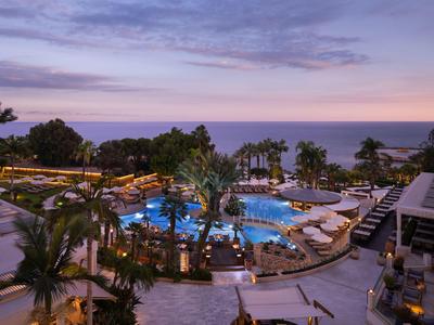 Abendaufnahme eines beleuchteten Hotelschwimmbeckens mit Palmen und Blick auf das Meer.