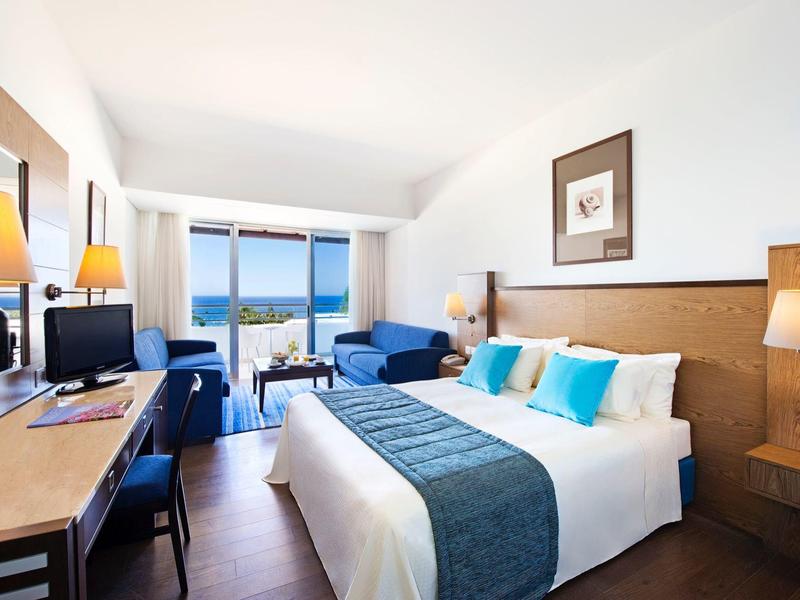 Bright hotel room with large bed, desk, and balcony overlooking the sea.