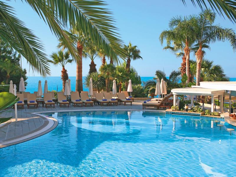 Hotel pool with palm trees, sun loungers, and sea view under clear sky.