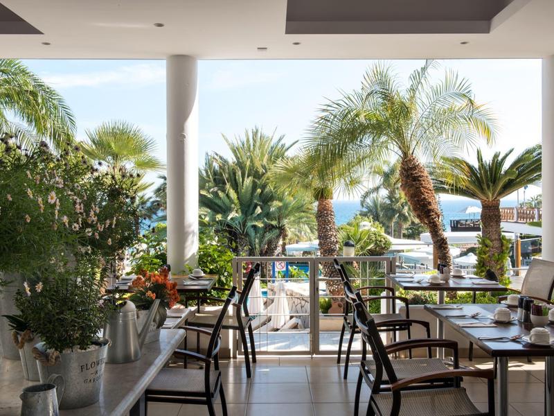 Restaurant terrace with chairs and tables, view of palm trees and sea in the background.