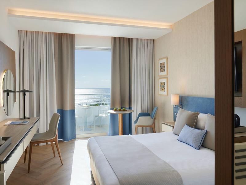Bright hotel room with double bed, desk, and sea view through panoramic glass.