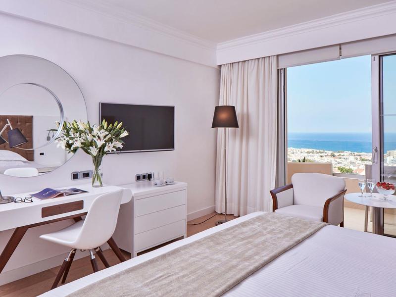 Modernes Hotelzimmer mit Schreibtisch, Fernseher, Sitzecke und Meerblick durch große Fenster.