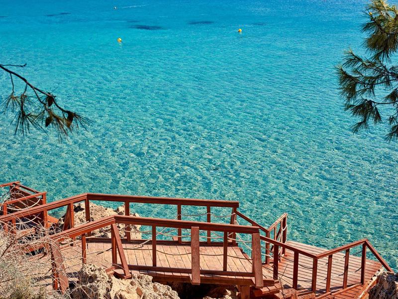 Holzstufen führen zu einem klaren, türkisfarbenen Meer mit Felsen im Vordergrund.