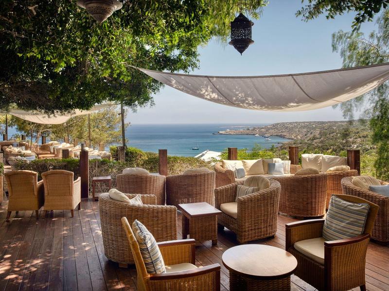 Gemütliche Außenterrasse mit Korbsesseln und Blick aufs Meer unter schattenspendenden Segeln.