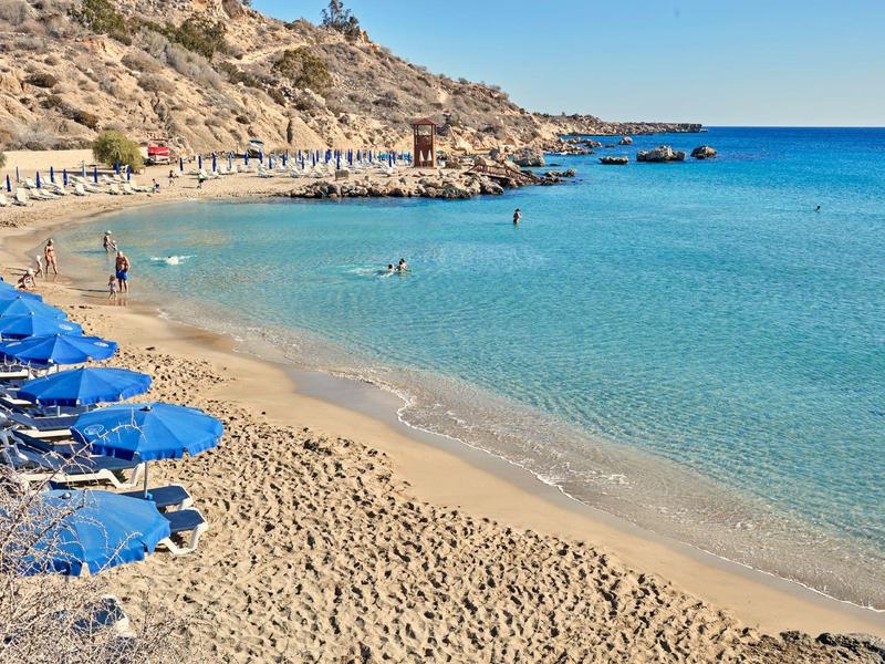 Sandstrand mit blauen Sonnenschirmen, klarem Wasser und felsigem Hintergrund.