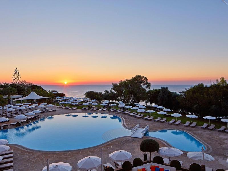 Schwimmbad mit Liegestühlen und Sonnenuntergang am Meer im Hintergrund