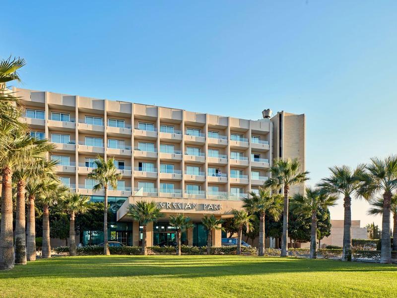 Ein mehrstöckiges Hotel mit Balkonen, umgeben von Palmen und einer grünen Rasenfläche bei klarem Himmel.