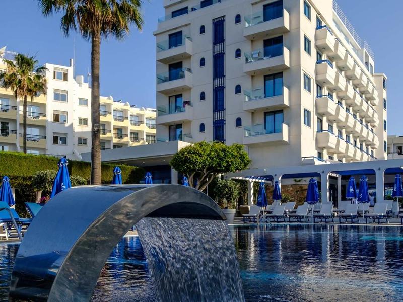 Moderner Hotelbau mit Pool, Wasserspiel und Palmen unter klarem blauem Himmel.