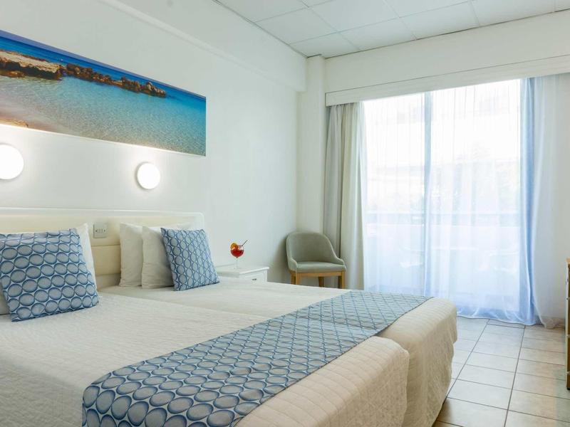 Helles Schlafzimmer mit zwei Einzelbetten, blauen Kissen, einem Sessel und großem Fenster mit Vorhängen.