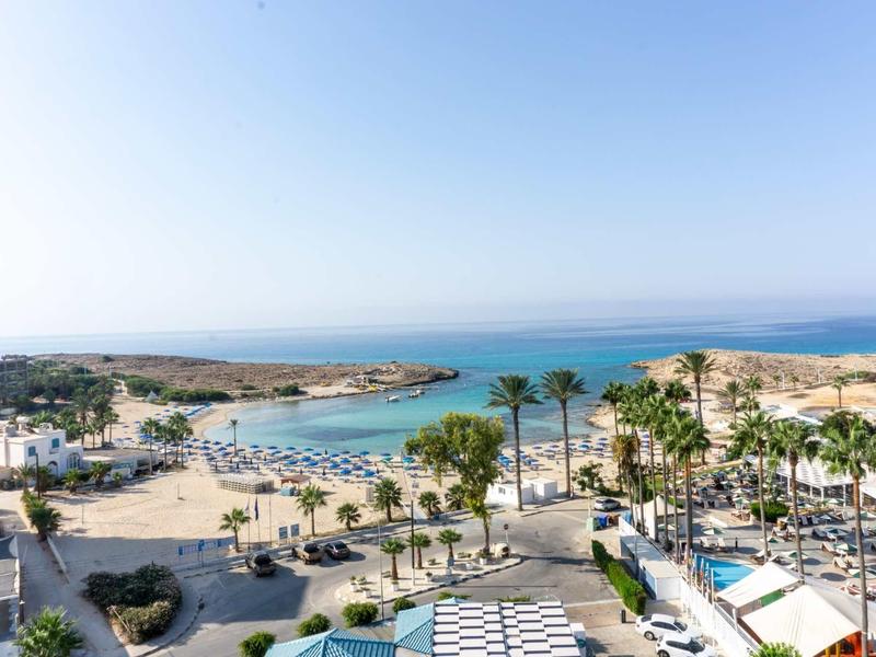 Küste mit blauem Meer, Sandstrand, Palmen und weißen Gebäuden unter klarem Himmel.