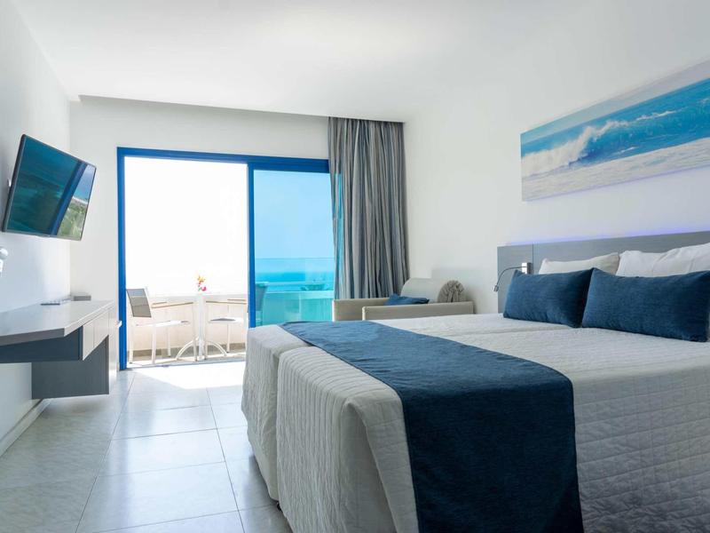 Helles Hotelzimmer mit großem Bett, blauen Akzenten, Flachbildschirm und Blick auf das Meer.