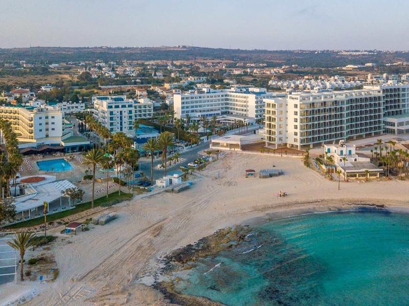 Strand mit Sand, klarem blauem Wasser und Hotels in mehreren Etagen im Hintergrund.