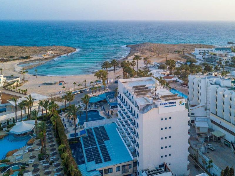 Blick auf ein Hotelresort am Meer mit Pools, weißen Gebäuden und einer Küstenlinie im Hintergrund.