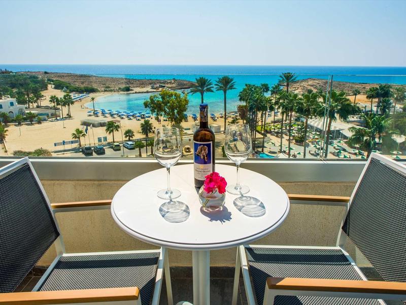 Balkontisch mit zwei Gläsern, Bierflasche und Blume, Blick auf Palmen und blaues Meer.