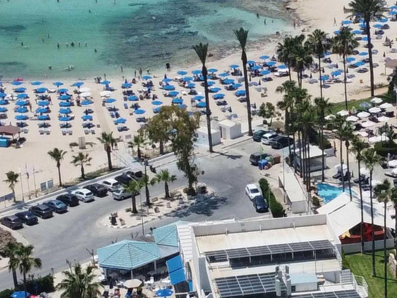 Strand mit blauen Sonnenschirmen, türkisfarbenem Wasser und angrenzendem Hotelkomplex.