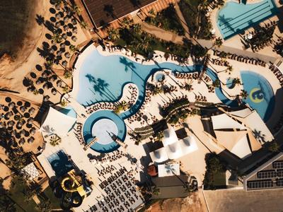 Vue aérienne d'une grande piscine d'hôtel avec chaises longues, palmiers et toboggans aquatiques.