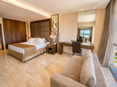 Chambre d'hôtel lumineuse avec lit double, coin canapé et vue sur la ville à travers de grandes fenêtres.
