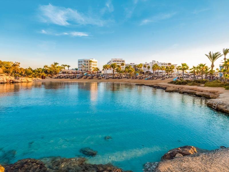 Klares blaues Wasser in einer Bucht mit Hotelgebäuden und Palmen im Hintergrund.
