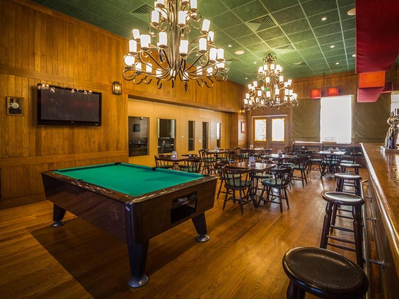 Ein großer Raum mit Billardtisch, Holzwänden, Tischen und Stühlen, beleuchtet durch Kronleuchter.