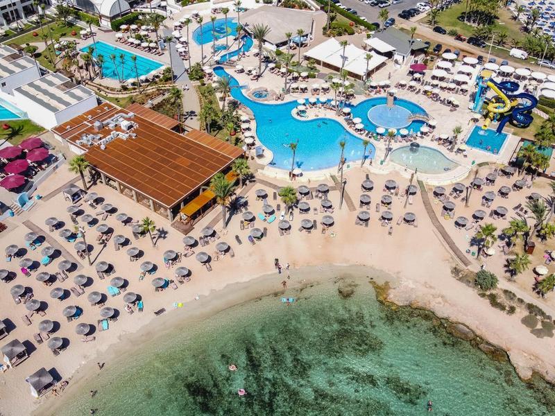 Luftaufnahme eines Strandes mit Liegestühlen, Sonnenschirmen und einem großen Resort-Poolbereich.