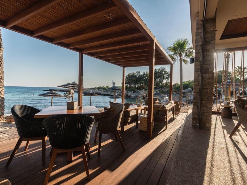 Terrasse mit Stühlen und Tischen mit Blick auf das Meer und Palmen bei Sonnenschein.