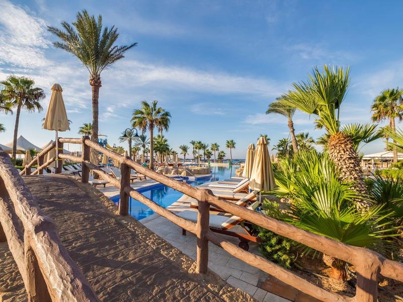 Ein sonniger Hotelpoolbereich mit Palmen, Holzgeländer und blauen Liegen unter blauem Himmel.