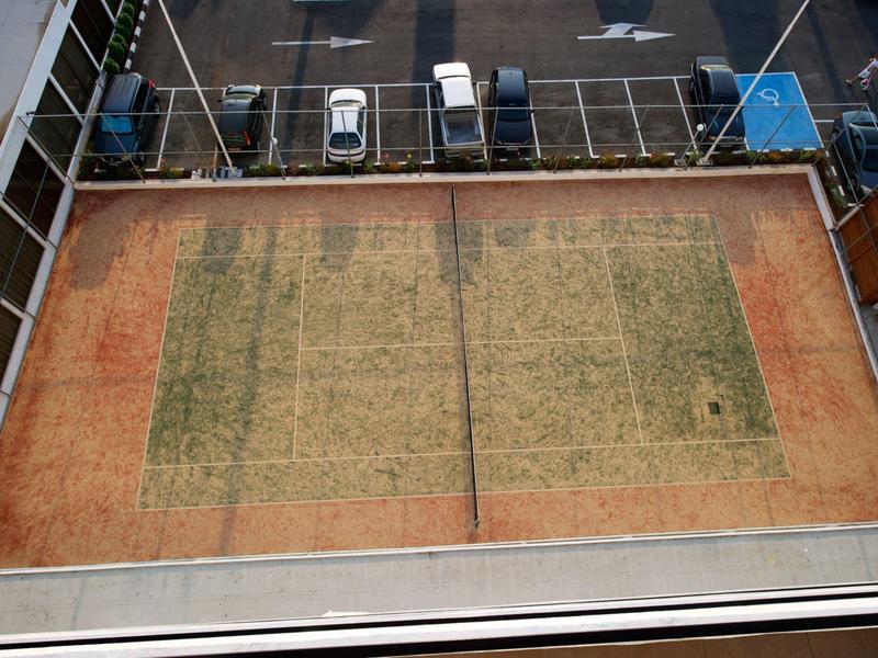 Abgenutztes Tennisfeld mit verblasster Linienmarkierung und umliegendem Parkplatz.