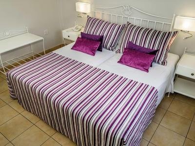 Doppelbett mit gestreifter Bettwäsche in Weiß, Lila und Pink, daneben weiße Nachttische mit Lampen.