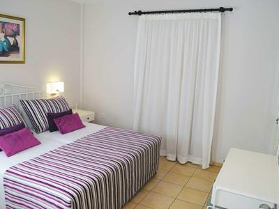 Schlafzimmer mit Doppelbett, gestreifter Bettwäsche, weißen Gardinen und einem Nachttisch mit Lampe.