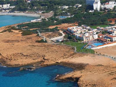 Küstenlandschaft mit felsigem Ufer, blauem Meer und Siedlung mit Häusern und Schwimmbad im Hintergrund.