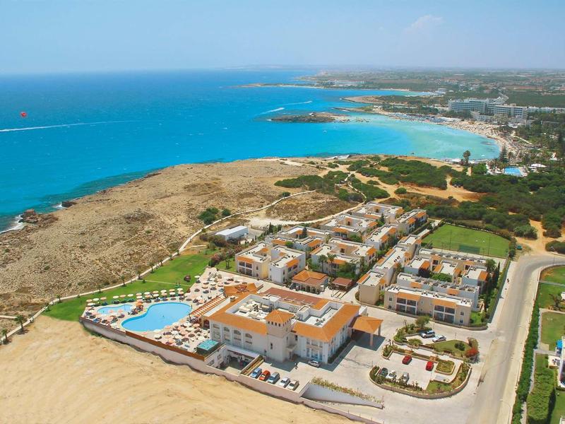 Luftaufnahme einer Küstenanlage mit Hotel, Pool, Sandstrand und klarem blauem Meer unter hellem Himmel.